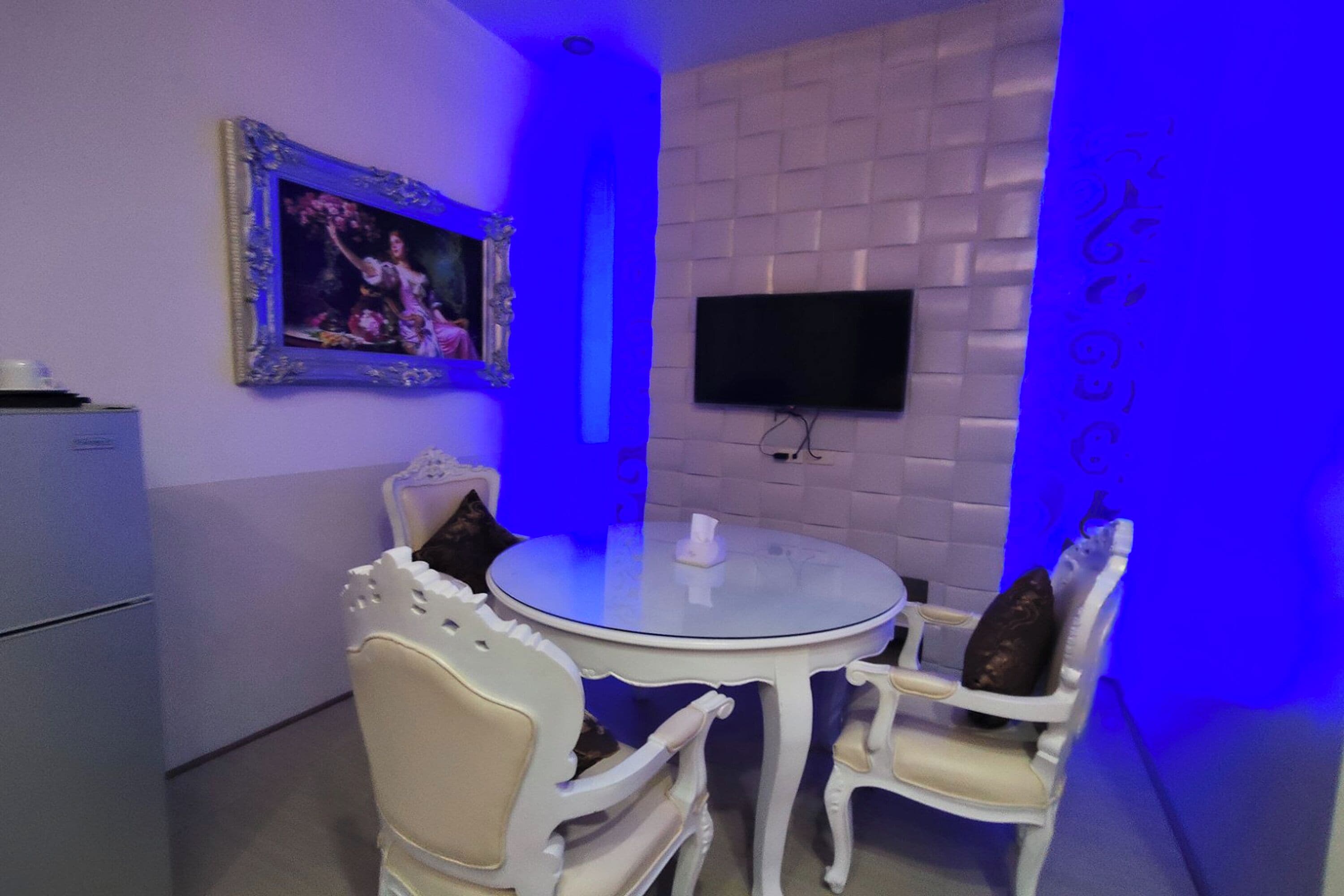 Una habitación con una mesa redonda y dos sillas, un televisor de pantalla plana montado en la pared, y un cuadro enmarcado sobre la mesa.