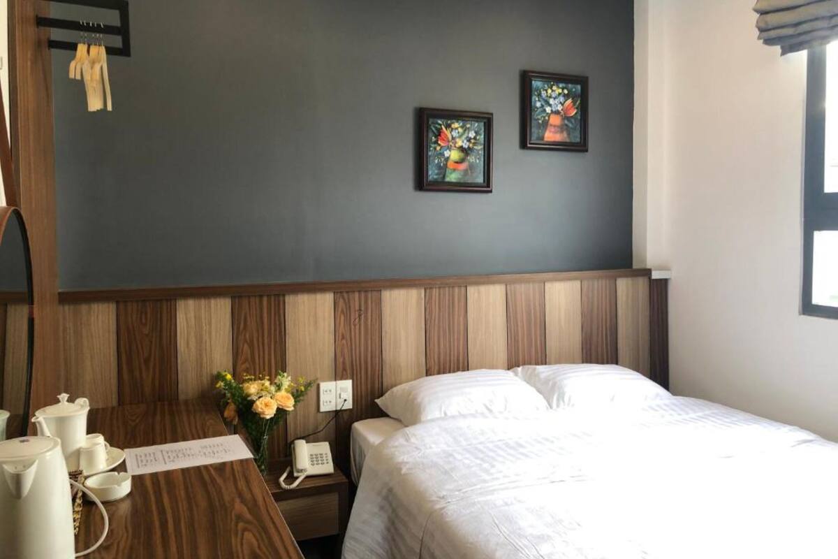 A hotel room with a wooden headboard, a bed with white linens, a bedside table with a phone and a vase of flowers, and two framed pictures on the wall.