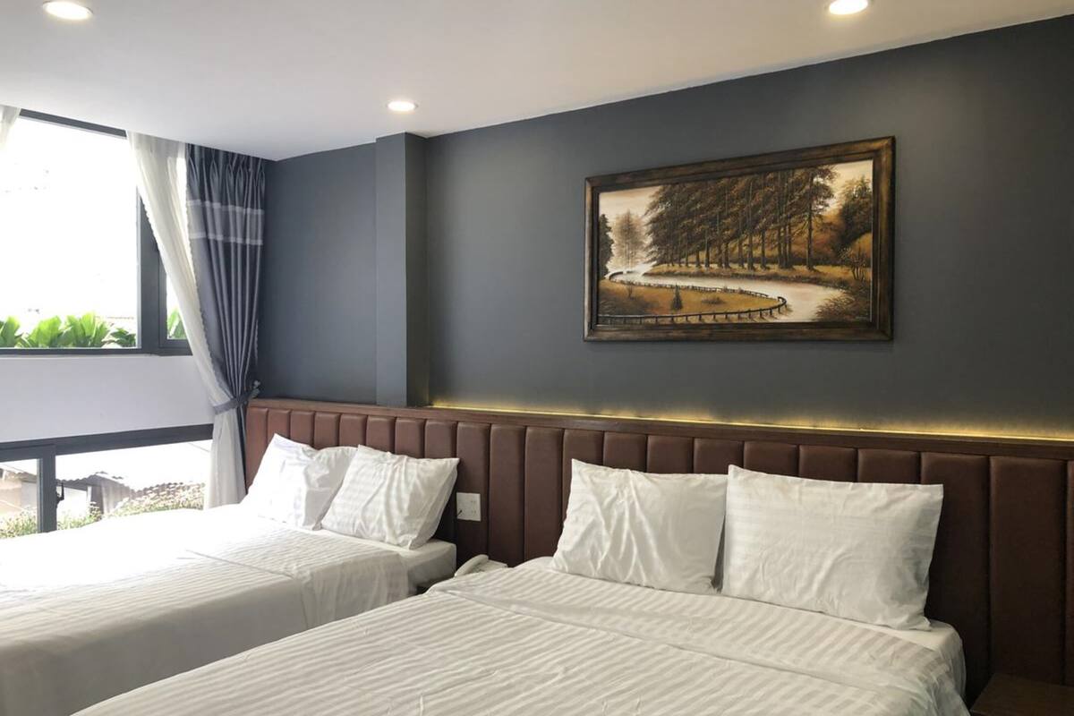 A hotel room with two beds, a large window with curtains, a painting on the wall, and a view of greenery outside.