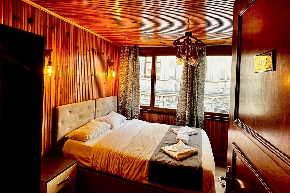 A wooden-paneled room with a large bed, a chandelier, and a window with curtains.