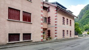 Un edificio rosa con contraventanas rojas, un balcón y un paisaje montañoso de fondo.