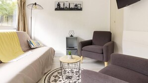 Una habitación moderna y bien iluminada con sofá, sillón y una mesita auxiliar. Un televisor de pantalla plana está montado en la pared y hay una estantería con objetos decorativos.