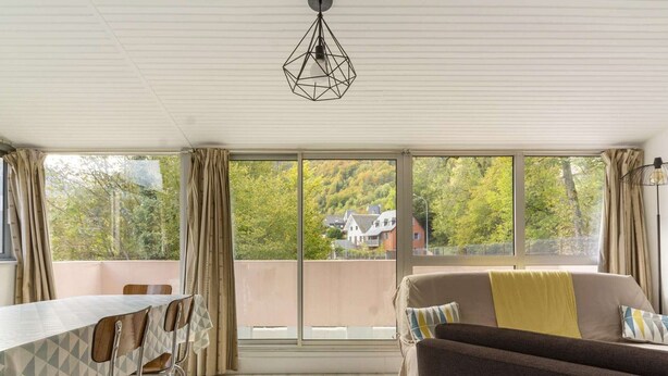 Una habitación con una mesa de comedor, sillas, un sofá y ventanas amplias que dan a vistas de árboles y casas.