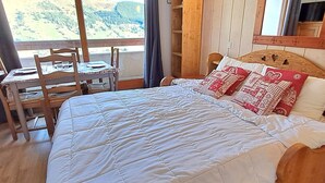 A bedroom with a bed, a wooden headboard, a bedside table, a chair, and a window with a view of a mountainous landscape. - Calm T2 apartment, slopes access, private parking (Les Deux Alpes)