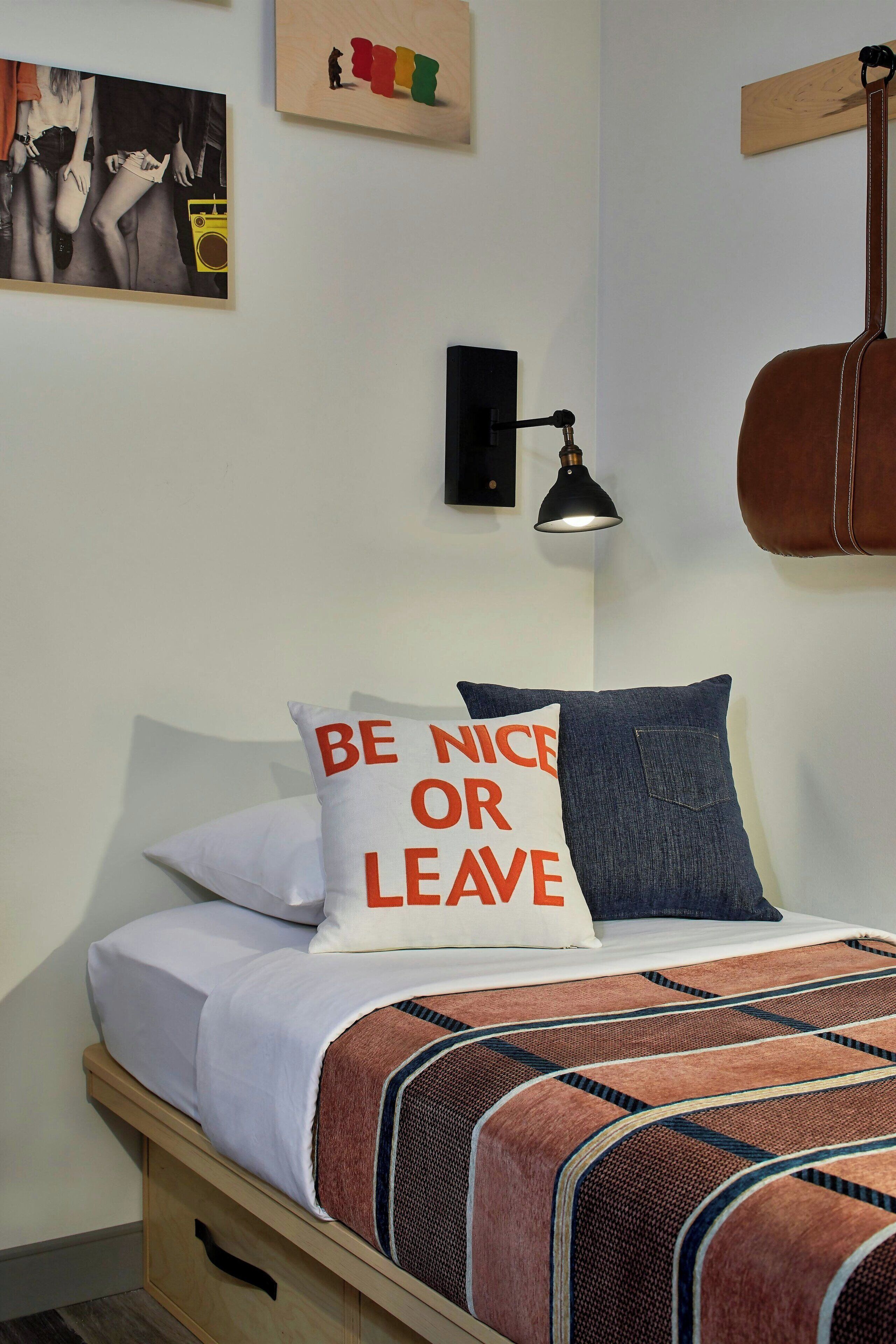 A neatly made bed with a patterned blanket, two pillows with text, a wall-mounted lamp, and a guitar hanging on the wall.