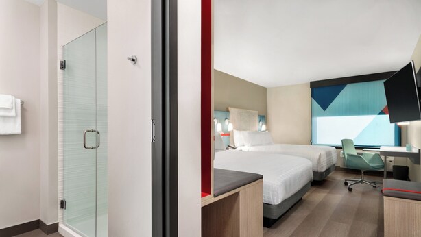 A hotel room with a glass shower enclosure, a bed with a red accent, a desk with a chair, and a large abstract painting on the wall. - avid hotels Perry-National Fairground Area by IHG (Perry)