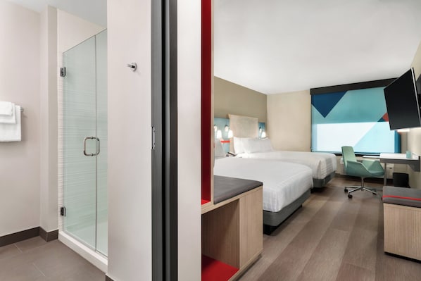 A hotel room with a glass shower enclosure, a bed with a red accent, a desk with a chair, and a large abstract painting on the wall. - avid hotels Perry-National Fairground Area by IHG (Perry)