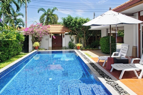 A swimming pool with tiled edges, surrounded by potted plants and a patio area with white chairs and a table.