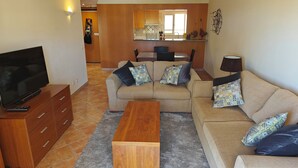 A living room with a sofa, a coffee table, a TV on a wooden cabinet, and a kitchen in the background. - Clube Alvor Ria 103 Luxury sea view 1bed apartment outstanding central location. (Alvor)