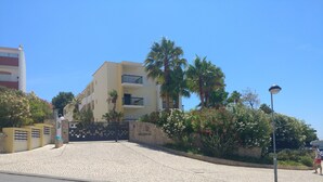 A multi-story building with balconies, a paved entrance area, and a stone wall with flowering plants. - Clube Alvor Ria 103 Luxury sea view 1bed apartment outstanding central location. (Alvor)