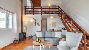 A spacious living area with a grey sofa, a white armchair, and a wooden coffee table. A wooden staircase leads to an upper level. - Casas Hortênsia - Oceanfront Home w/ Sunset Views (Mosteiros)