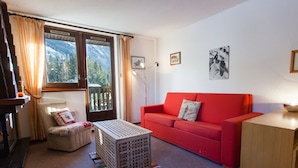A cozy living room with a red sofa, a wooden cabinet, a small table, and a chair. There are two windows with curtains, a radiator, and a framed picture on the wall.