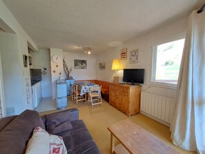 A living room with a sofa, a wooden table, a dining area with chairs, a TV, and a window with curtains.