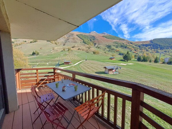 A balcony with a table and chairs, overlooking a green landscape with hills and a clear sky.