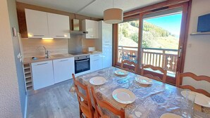 Une cuisine moderne avec des meubles blancs, une table à manger pour quatre personnes et une vue sur un paysage montagneux par le biais d’une grande fenêtre.