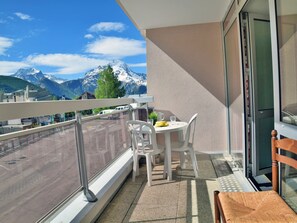 En balkon med et bord og stole, udsigt til et bjerglandskab, og en bygning med teksten "Le Meilleur" i baggrunden.