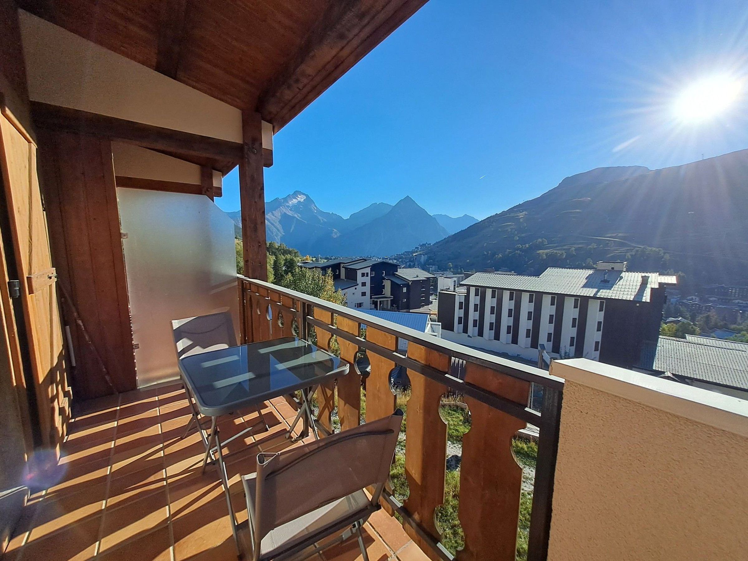 Un balcon avec une table et une chaise, donnant sur un paysage montagneux avec des bâtiments visibles au loin.