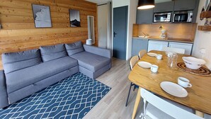 Un espace de vie moderne comprenant un canapé d’angle gris, une table à manger en bois mise avec des assiettes et des tasses, et un coin cuisine équipé d’un four à micro-ondes et de placards.