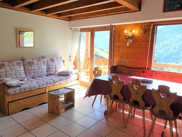 A cozy room with a wooden sofa, a dining table with heart-shaped chairs, and a view of the outdoors.