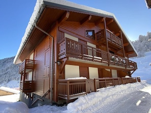 A wooden chalet with a balcony, a covered entrance, and a snow-covered landscape.
