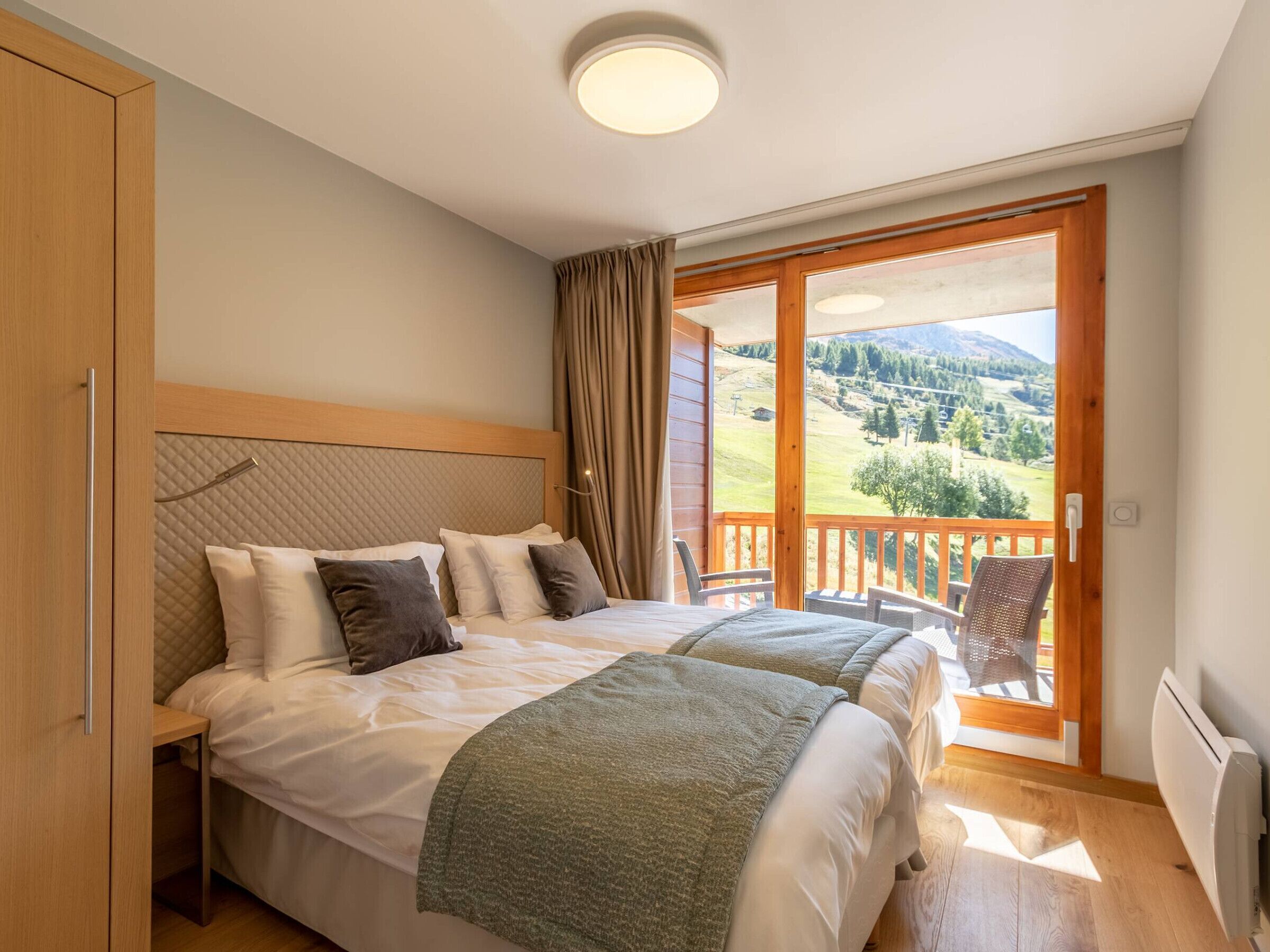 Une chambre avec deux lits, une tête de lit en bois, une vue sur un paysage montagneux par une grande fenêtre et un balcon agrémenté de meubles en osier.
