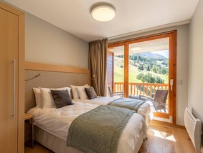 A bedroom with two beds, a wooden headboard, a view of a mountainous landscape through a large window, and a balcony with wicker furniture.