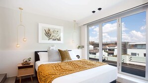 A modern bedroom with a large bed, a nightstand, a framed picture, and a view of the city through large windows. - Perfect Located 2-Bed with Spacious Balcony & BBQ (Cremorne)