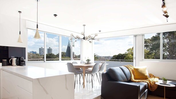 A modern kitchen with a large island, dining area with chairs, and a sofa with a view of the city. - Perfect Located 2-Bed with Spacious Balcony & BBQ (Cremorne)