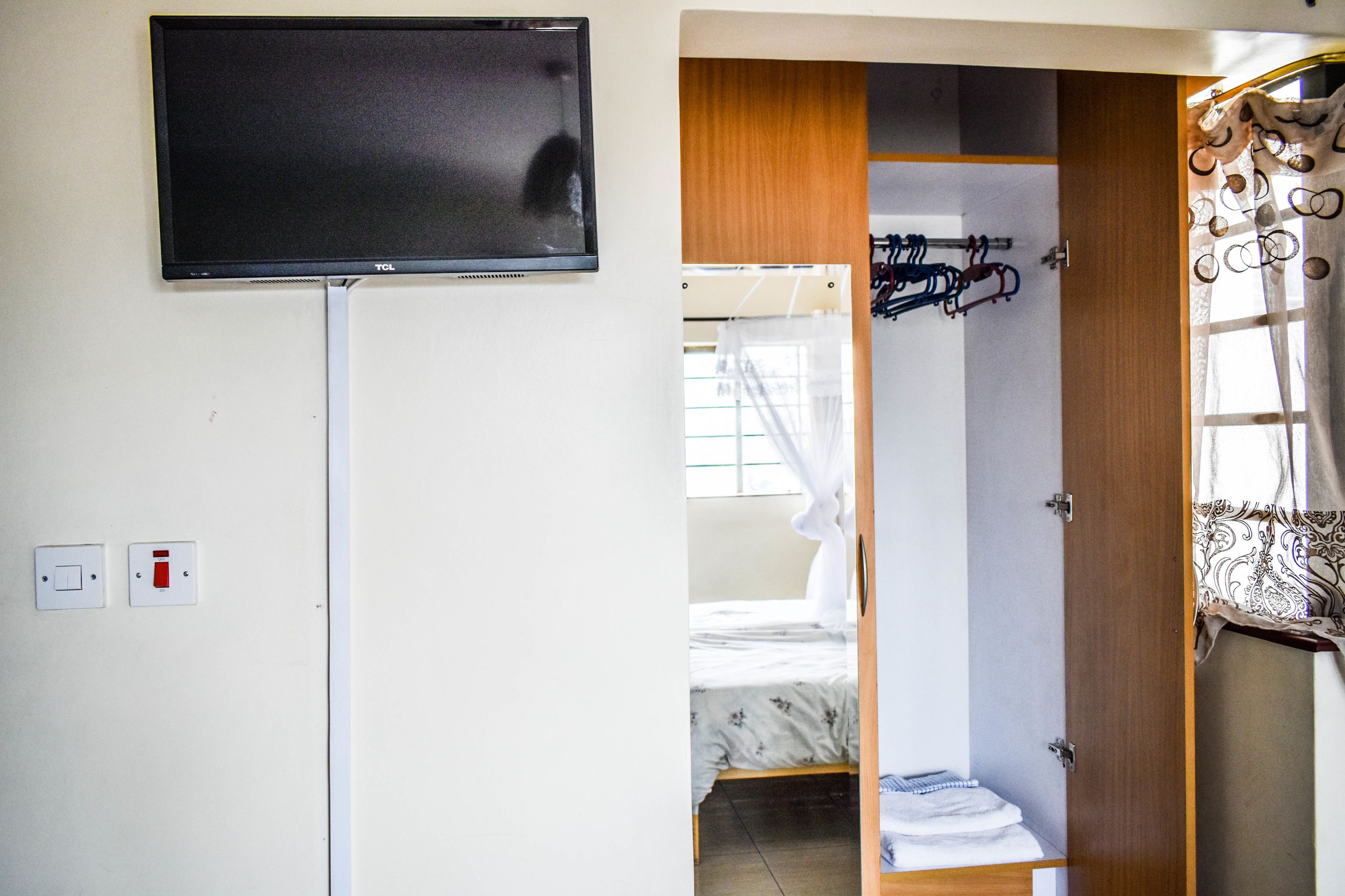 A room with a mounted television, a bed with a floral bedspread, a wooden wardrobe, and a window with a curtain.