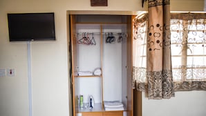 A room with a wardrobe containing hanging clothes, a flat-screen TV mounted on the wall, and a window with patterned curtains. - Affordable furnished studio apartment (Nairobi)