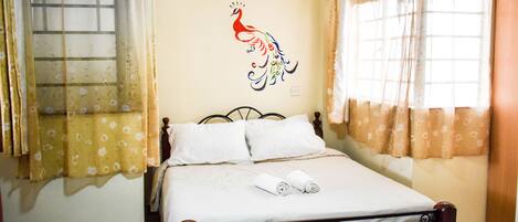 A neatly made bed with white linens and rolled towels, a wall with a motivational quote and a peacock illustration, and a window with curtains.