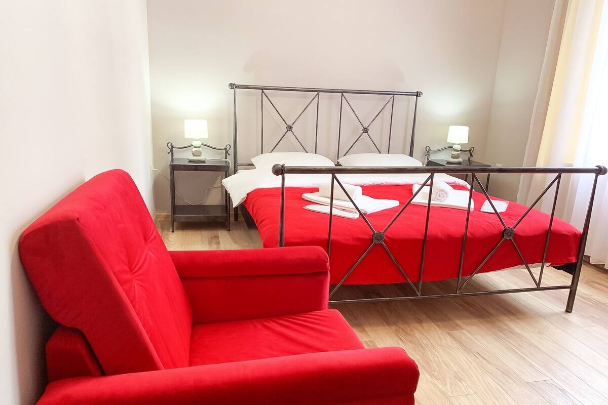 Una habitación con una cama roja, un sillón rojo, una cama con estructura de metal, dos mesitas de noche con lámparas y suelo de madera.