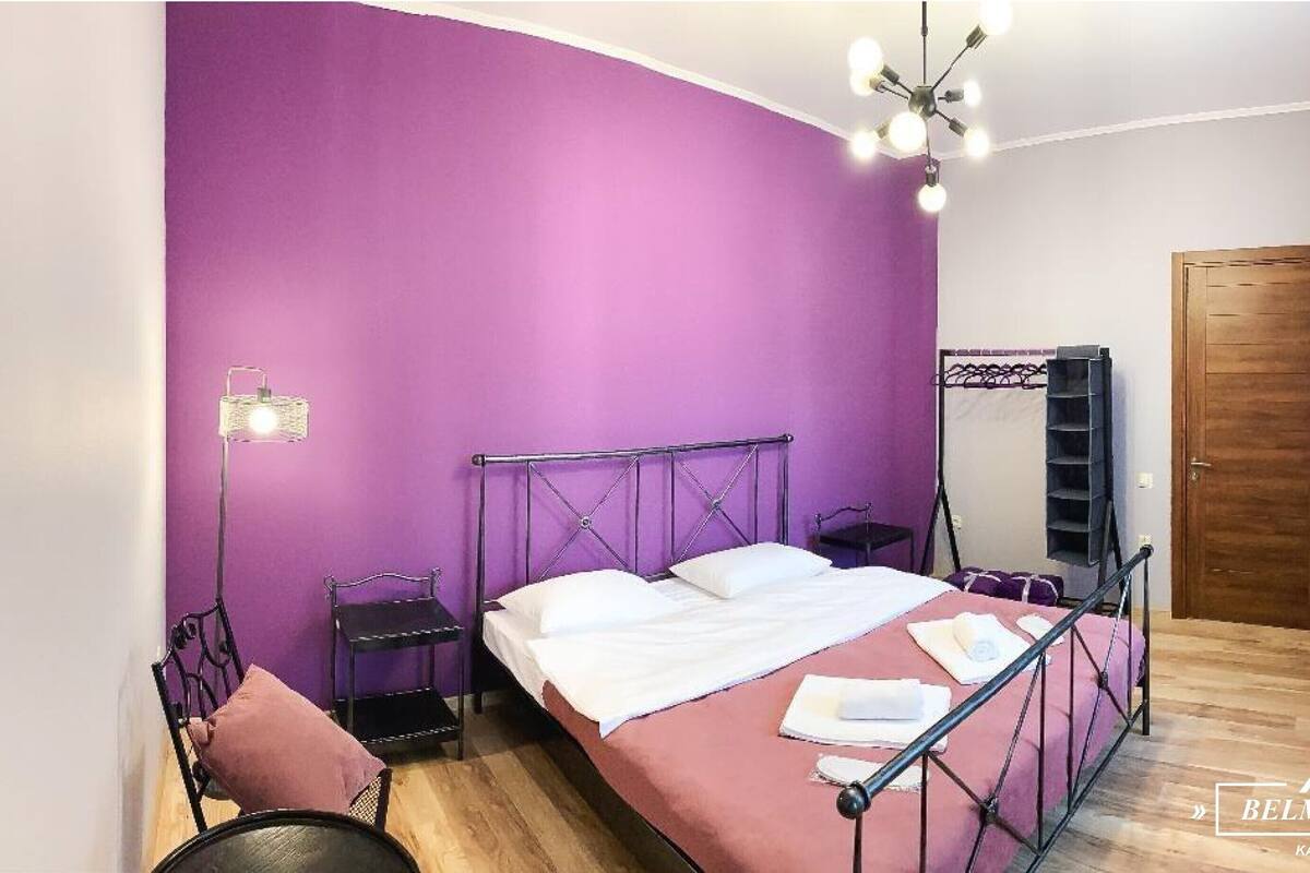 Un dormitorio con pared morada, estructura de cama metálica, mesita de noche, estante colgante y puerta de madera.