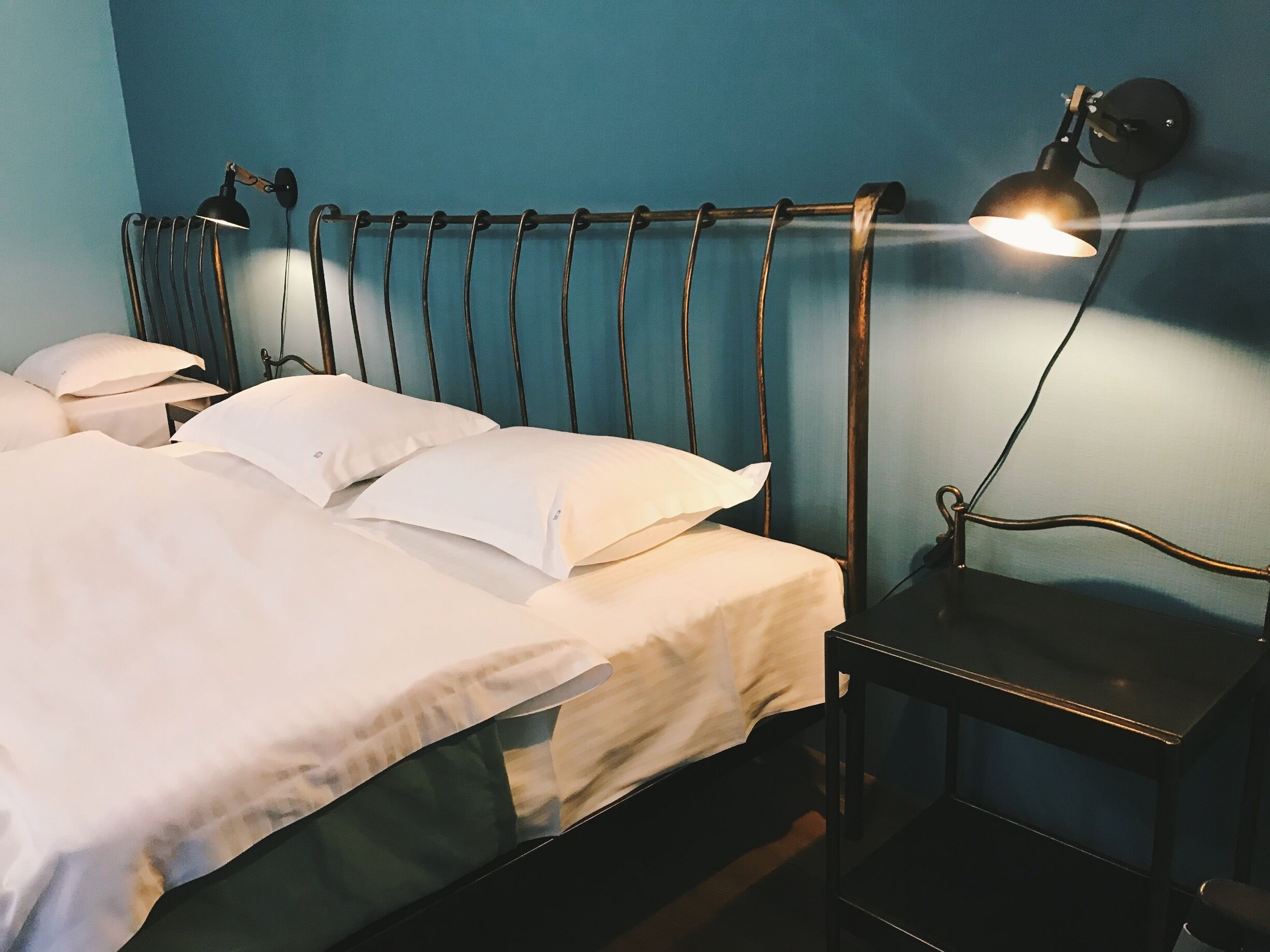 Ein ordentlich bezogenes Bett mit weißen Leinen und einem Metall-Kopfteil, ein Nachttisch mit Lampe und eine blaue Wand.