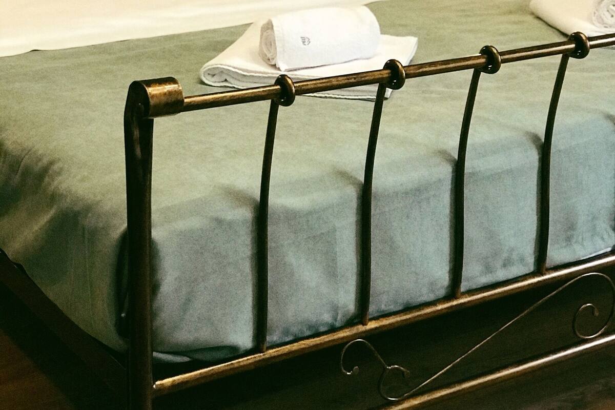 Una cama bien hecha con cabecera de metal, una lámpara de noche y una toalla doblada sobre la cama.
