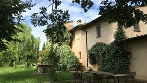 Un espace de restauration extérieur rustique, avec un banc en pierre et des chaises en métal, entouré d’une végétation luxuriante et bordé d’un bâtiment doté d’un toit en tuiles.