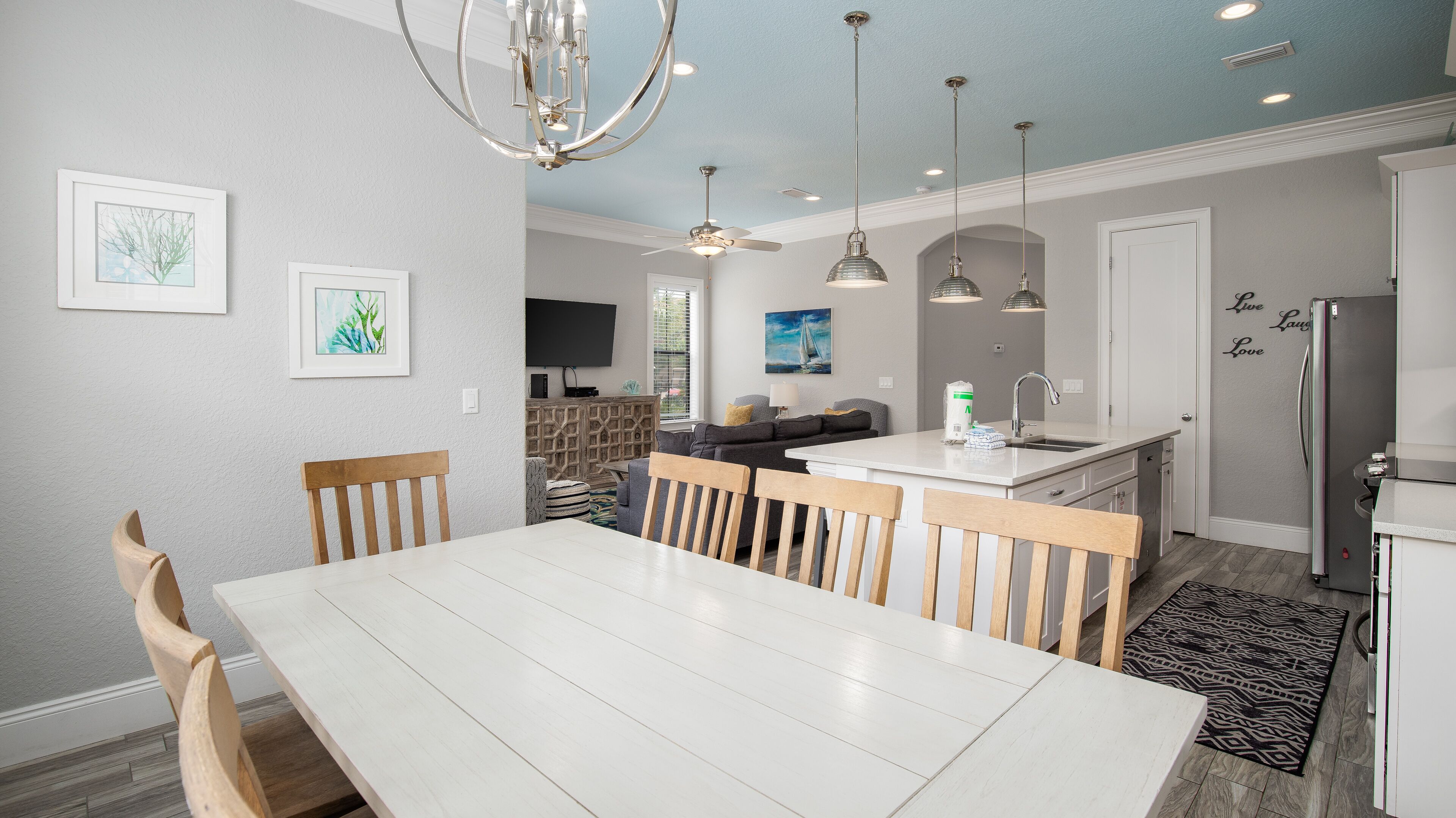 A modern kitchen with a central island, a dining area with a white table and wooden chairs, and a living area with a sofa and a TV.