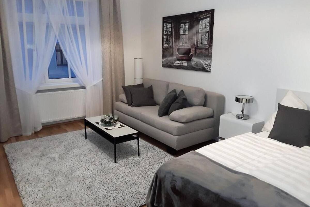 A modern hotel room with a grey sofa, a small table, a bed with a white and grey duvet, a wall-mounted picture, and a window with sheer curtains.