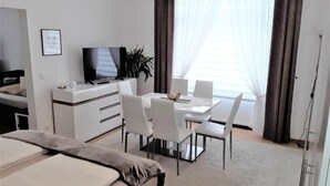 Quarto de hotel com cama, secretária com televisão, mesa de jantar com cadeiras e uma grande janela com cortinas.