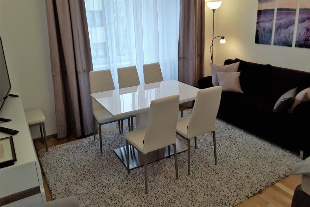A modern living room with a dining area, featuring a white table and chairs, a black sofa, and a large window with curtains.