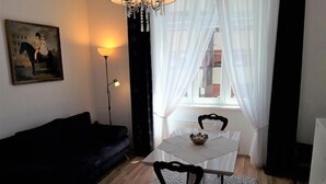 Um quarto com um sofá preto, uma mesa branca com duas cadeiras, um candelabro e um quadro na parede.