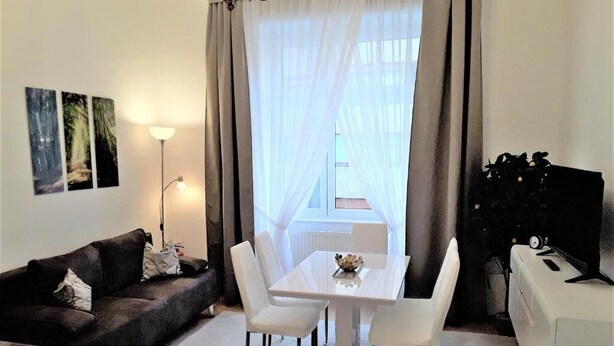 Sala de estar com sofá escuro, mesa de jantar branca com quatro cadeiras, abajur e radiador.