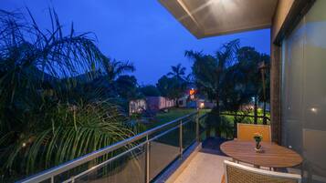 A balcony with a glass railing overlooking a garden with palm trees and a view of the cityscape at dusk.