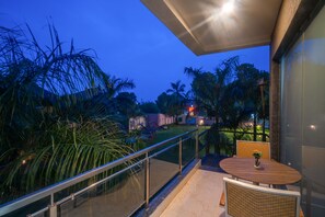 A balcony with a glass railing overlooking a garden with palm trees and a view of the cityscape at dusk. - Hotel Natraj & Resort (Mansa)