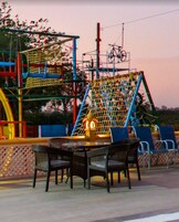 Un área de estar al aire libre con sillas de metal y una mesa, rodeada por un parque infantil con redes de escalada y un tobogán.