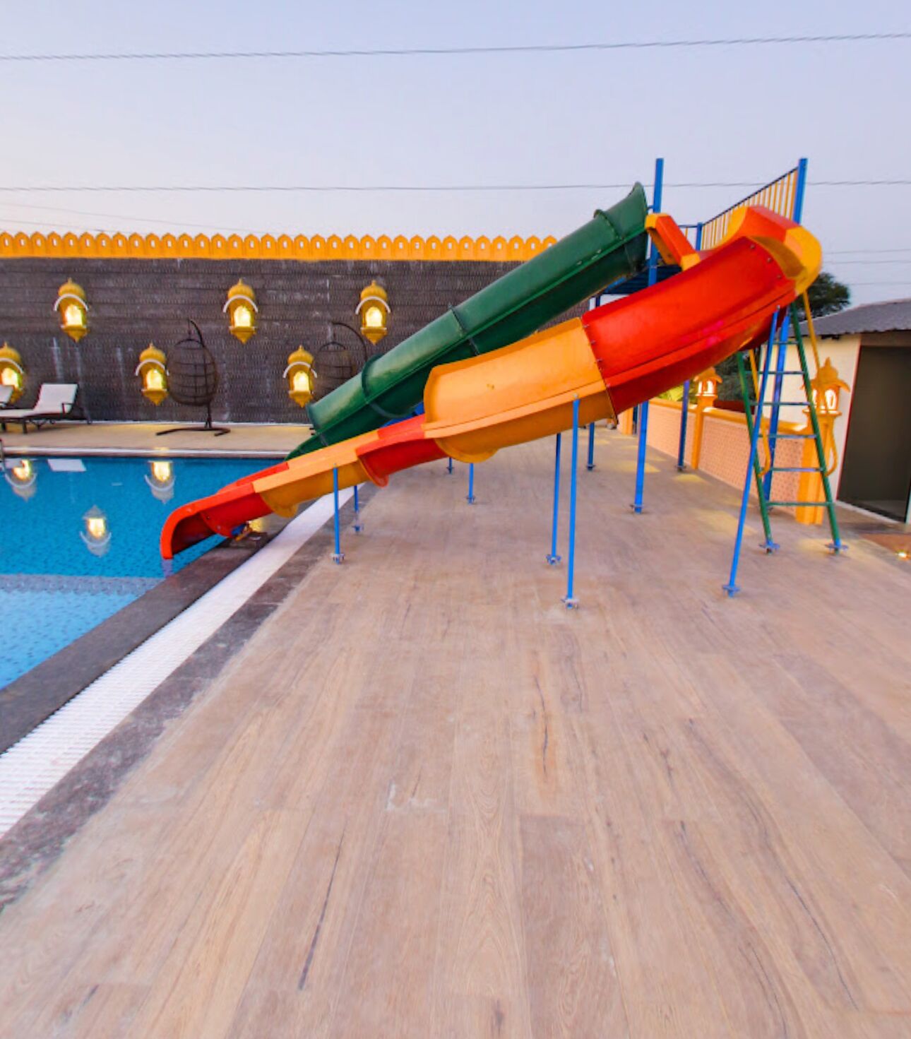 A multi-colored slide structure next to a swimming pool with a wooden deck and decorative lights.