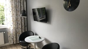 Ett modernt rum med en platt-TV som är monterad på väggen, en rund svart klocka och två svarta stolar runt ett litet vitt bord.