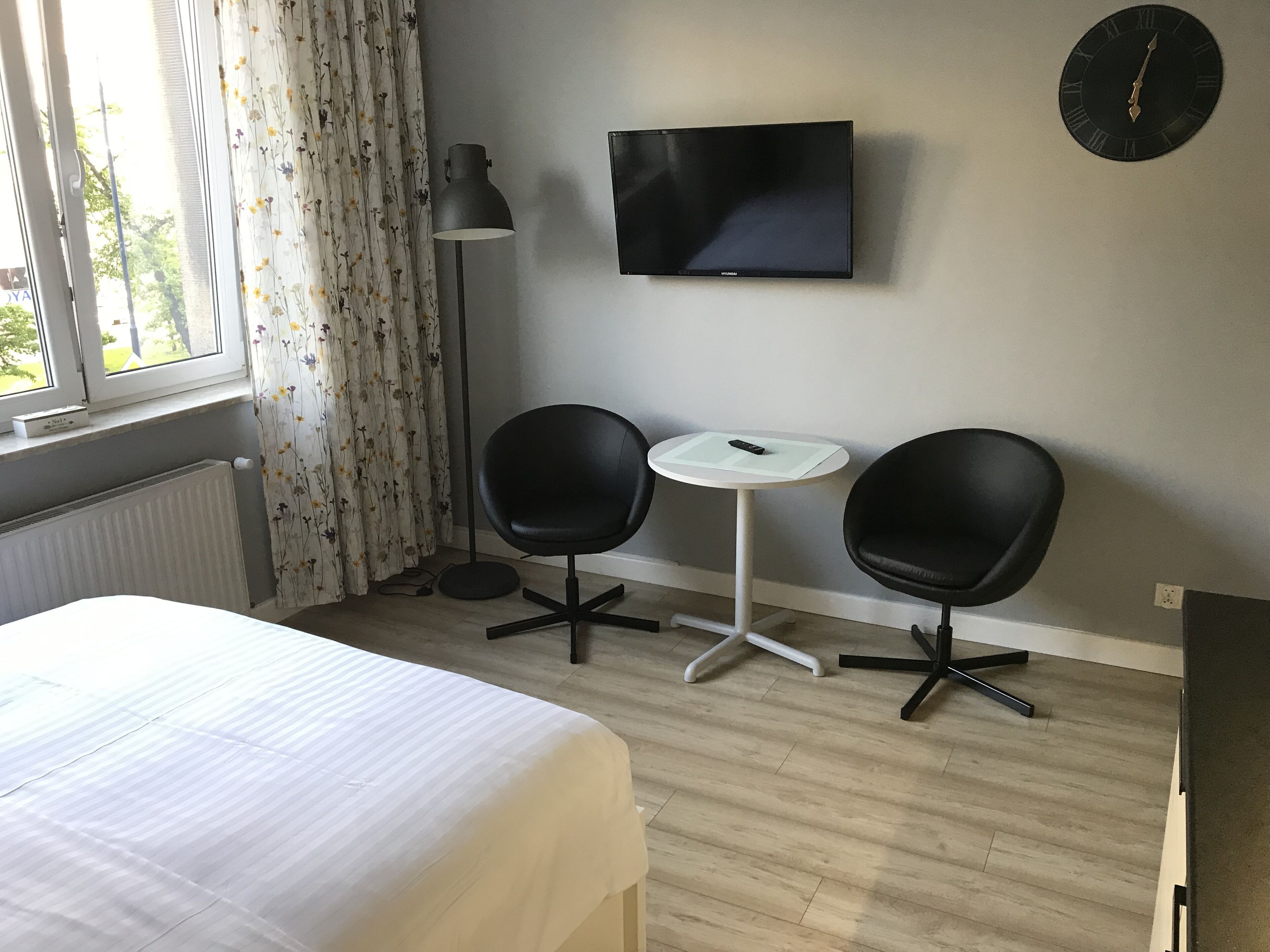 Une chambre d’hôtel avec un lit, deux chaises noires, une petite table blanche, un téléviseur et une horloge au mur.