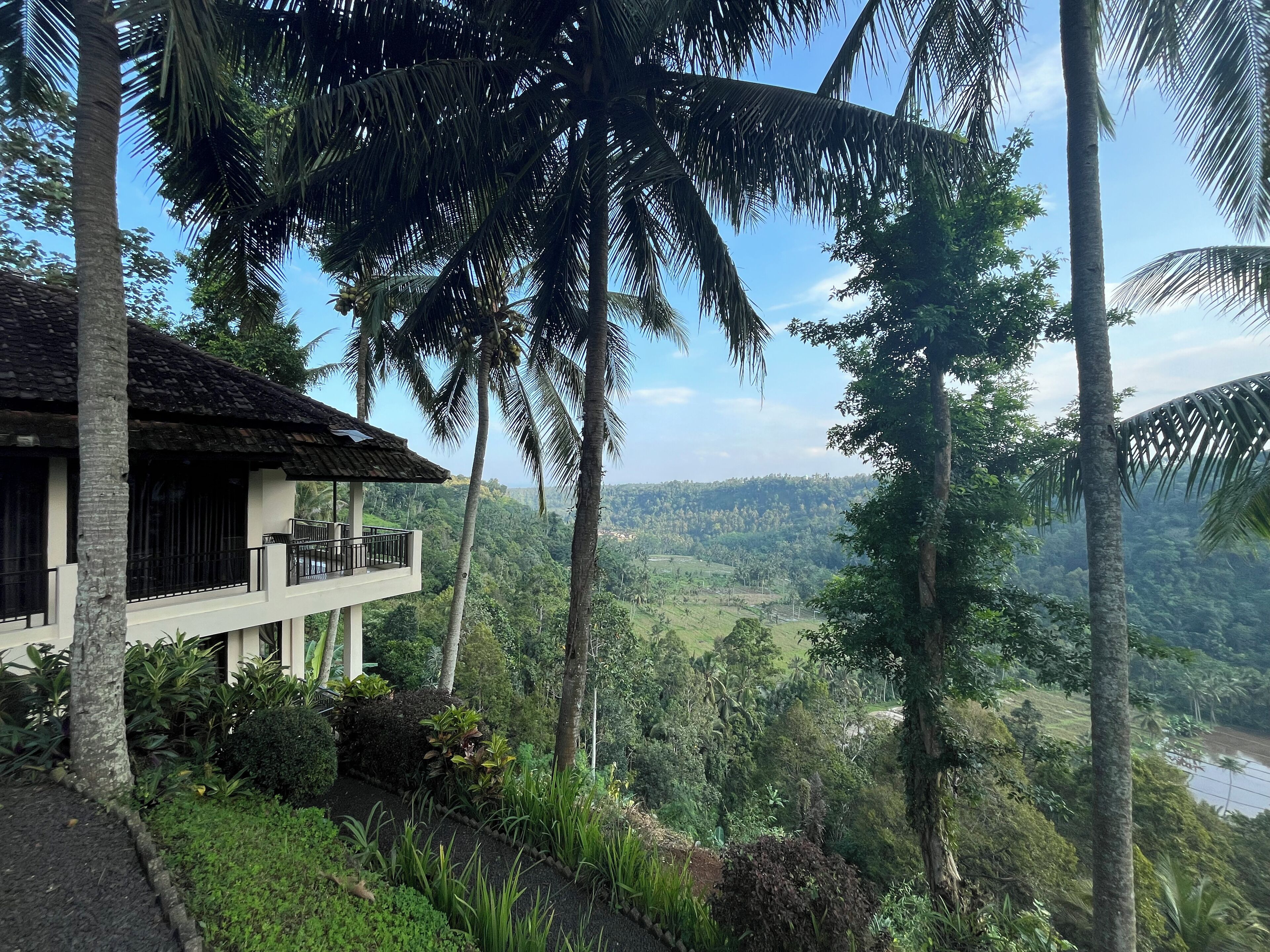 Un edificio de dos pisos con balcón, rodeado de exuberante vegetación y palmeras, con vista a un paisaje montañoso.