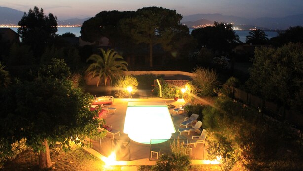 Una piscina con sillones de descanso y un jacuzzi, rodeada de árboles y vista a la ciudad al anochecer.